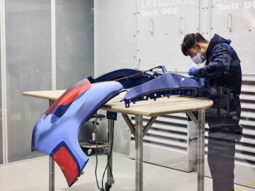 Een persoon die beschermende kleding draagt, werkt aan blauwe en rode carrosseriedelen van de BMW 3.0 CSL, geplaatst op een tafel in een industriële omgeving.