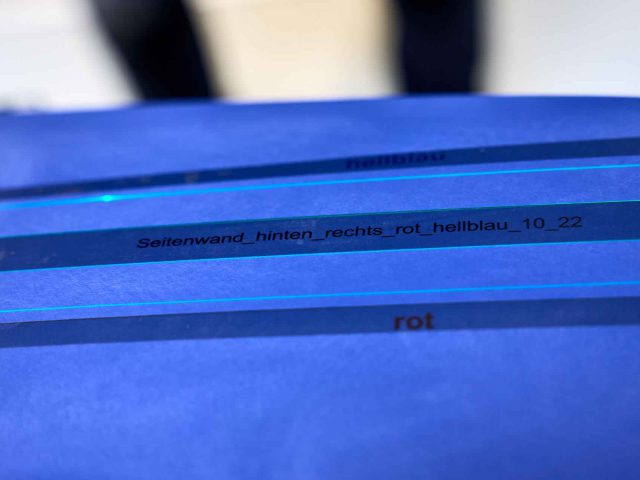 Close-up van tekstlabels op blauw materiaal, waarbij op het bovenste label "hellblau" staat en op het onderste label "Seitenwand_hinten_rechts_rot_hellblau_10_22" en "rot", wat lijkt op de gedetailleerde labels die vaak te zien zijn op vintage BMW 3.0 CSL-modellen.