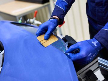 Een persoon die blauwe handschoenen draagt, brengt met een plat stuk gereedschap nauwgezet een sticker op een blauw oppervlak aan. De scène, die doet denken aan precisiewerk dat vaak wordt gezien bij het maken van voertuigen als de BMW 3.0 CSL, lijkt zich af te spelen in een werkplaats of productieomgeving.