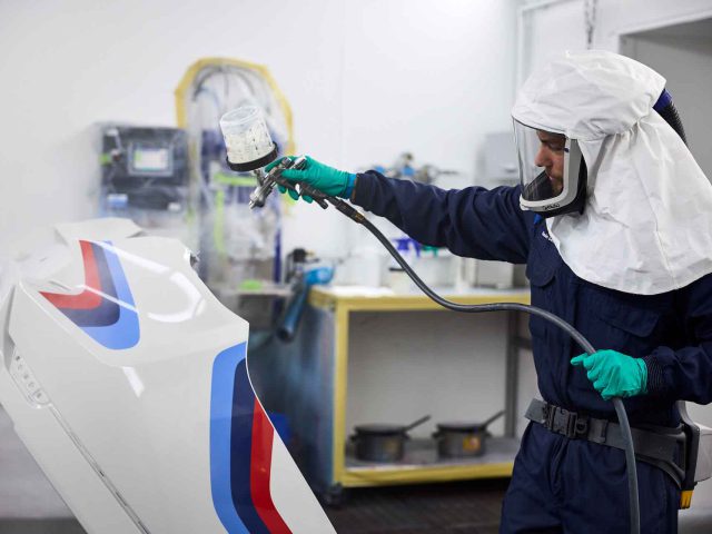 Een persoon die beschermende kleding en handschoenen draagt, spuit in een werkplaats een groot wit BMW 3.0 CSL-voertuigonderdeel met gekleurde strepen.