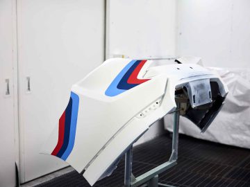 In een werkplaats wordt een witte BMW 3.0 CSL-bumper met blauwe en rode strepen tentoongesteld, gemonteerd op een metalen standaard.