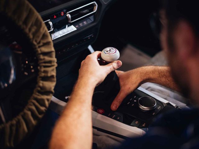 Persoon die een handmatige versnellingspook bedient in een BMW 3.0 CSL, met de rechterhand op de pookknop en de linkerhand bij de middenconsole.