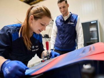 Twee technici in blauwe uniformen werken in een industriële omgeving aan een machine, vergelijkbaar met de precisie die je ziet bij het onderhouden van een BMW 3.0 CSL. De ene technicus bedient een stuk gereedschap, terwijl de ander aandachtig observeert.