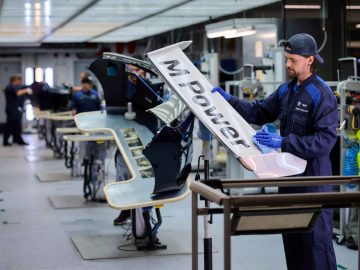 Een werknemer in een blauwe overall inspecteert een groot auto-onderdeel met het opschrift "M Power" in een productiefaciliteit, terwijl andere werknemers en apparatuur op de achtergrond zichtbaar zijn. De scène benadrukt het ingewikkelde vakmanschap achter de iconische BMW 3.0 CSL.