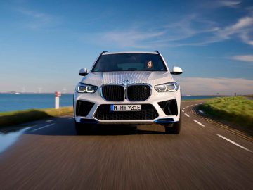 Een witte BMW iX5 Hydrogen SUV rijdt onder een helderblauwe lucht over een bochtige kustweg.