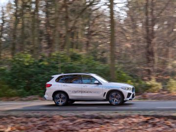 Een witte BMW iX5 Hydrogen SUV met op de zijkant 'Hydrogen' geschreven, rijdt over een weg door een bosrijke omgeving.