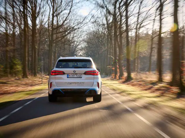 Een witte BMW iX5 Hydrogen SUV rijdt op een zonnige dag over een met bomen omzoomde weg in een bosrijke omgeving.