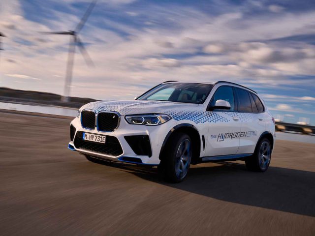 Een witte BMW iX5 Hydrogen SUV met het opschrift 'Hydrogen' rijdt op een weg met windturbines op de achtergrond onder een gedeeltelijk bewolkte hemel.