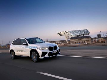 Een witte BMW iX5 Hydrogen SUV glijdt over een weg, omlijst door een modern, hoekig gebouw en een kraan op de achtergrond onder een heldere hemel.