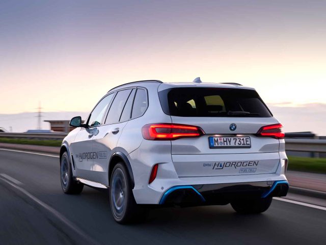 Een witte BMW iX5 waterstofbrandstofcelauto met een kentekenplaat met de tekst "M HY 731E" rijdt in de schemering over een weg.