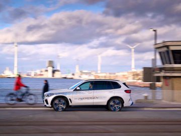 Een witte BMW iX5 Hydrogen SUV met de tekst "Hydrogen" op de zijkant rijdt langs een weg aan het water. Er passeert een fietser en op de achtergrond zijn windturbines zichtbaar.