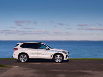 Een witte SUV met het opschrift "BMW iX5 Hydrogen Fuel Cell" rijdt op een weg naast een watermassa onder een heldere hemel.