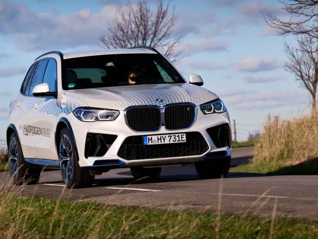 Een witte BMW iX5 Hydrogen SUV met een 'HYDROGEN FUEL CELL'-embleem rijdt over een weg omgeven door bomen en gras, allemaal onder een gedeeltelijk bewolkte hemel.