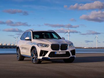 Een BMW iX5 Hydrogen SUV staat geparkeerd op een verhard terrein nabij een grote watermassa, met windturbines en een helderblauwe lucht bezaaid met verspreide wolken op de achtergrond.