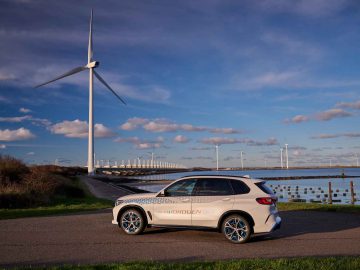 Een witte SUV met de tekst "BMW iX5 Hydrogen" staat geparkeerd op een weg vlakbij een grote windturbine; verschillende andere windturbines en een watermassa zijn zichtbaar op de achtergrond onder een blauwe lucht.