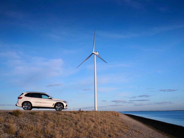 Een witte BMW iX5 Hydrogen SUV staat geparkeerd op een droge, met gras begroeide heuvel naast een grote windturbine met een watermassa op de achtergrond onder een helderblauwe lucht.