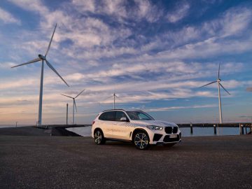 Een witte BMW iX5 Hydrogen SUV staat geparkeerd op een open plek vlakbij een waterkant, met verschillende windturbines op de achtergrond onder een gedeeltelijk bewolkte hemel.