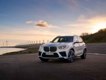 Een BMW iX5 Hydrogen SUV geparkeerd bij een watermassa bij zonsondergang met windturbines en een lange brug op de achtergrond.