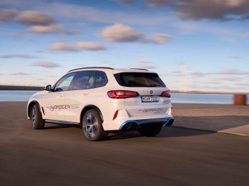 Een witte BMW iX5 Hydrogen SUV met het label 'Hydrogen Fuel Cell' rijdt op een verharde weg nabij een watermassa onder een gedeeltelijk bewolkte hemel.