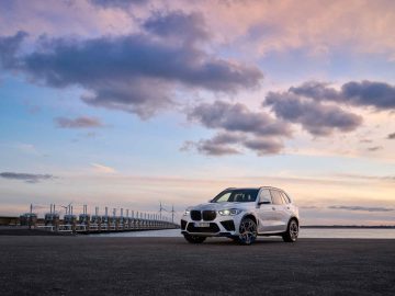 Een BMW iX5 Hydrogen staat geparkeerd op een open verhard terrein met een rij windturbines en een watermassa op de achtergrond, allemaal verlicht door een kleurrijke zonsonderganghemel.