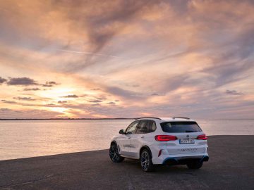 Een witte BMW iX5 Hydrogen SUV geparkeerd op een kustlijn, met een zonsondergang en een bewolkte lucht op de achtergrond. Op het kenteken van het voertuig staat "M.5987E.