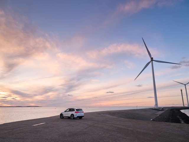 Een BMW iX5 Hydrogen staat bij zonsondergang geparkeerd aan de rand van een waterlichaam met twee grote windturbines op de achtergrond tegen een gedeeltelijk bewolkte hemel.