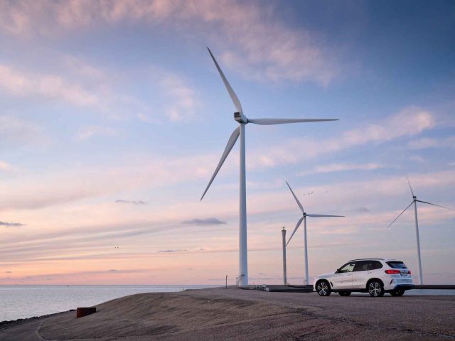 Een witte BMW iX5 Hydrogen staat bij zonsondergang geparkeerd bij drie grote windturbines aan de kust, onder een gedeeltelijk bewolkte hemel.