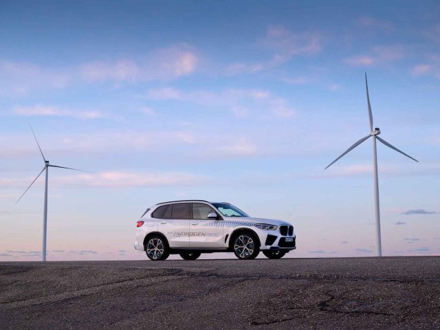 Een witte BMW iX5 Hydrogen SUV met een 'Hydrogen Fuel Cell'-logo staat geparkeerd op een weg, met twee windturbines hoog tegen de blauwe lucht op de achtergrond.