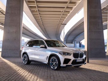 Een strakke, witte BMW iX5 Hydrogen staat geparkeerd onder een groot betonnen viaduct, waar zonlicht door de gaten in de constructie stroomt.