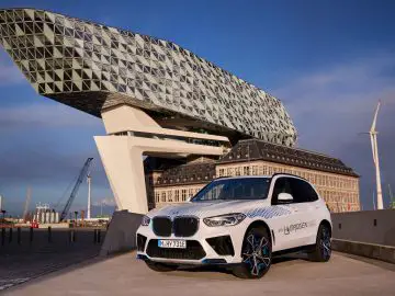 Een witte BMW iX5 Hydrogen SUV staat geparkeerd voor het Havenhuis in Antwerpen, België, onder een gedeeltelijk bewolkte hemel. Op de achtergrond is een windturbine zichtbaar.