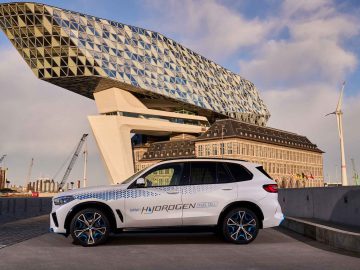 Een witte BMW iX5 Hydrogen SUV met de tekst 'Hydrogen Fuel Cell' staat op een heldere dag geparkeerd voor een modern, geometrisch gebouw.