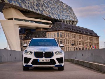 Vooraanzicht van een witte BMW iX5 Hydrogen geparkeerd op een weg met een modern, hoekig gebouw en historische structuur op de achtergrond. Aan de rechterkant zijn windturbines zichtbaar.