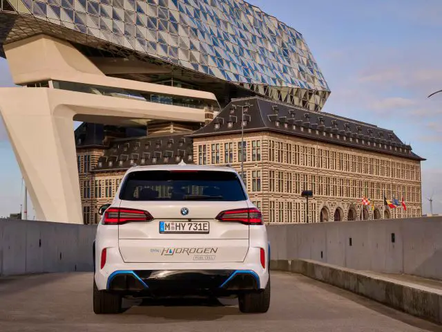 Een wit BMW iX5 waterstofbrandstofcelvoertuig met "HYDROGEN" op de achterkant, geparkeerd in een stedelijke omgeving met moderne en klassieke architectuur op de achtergrond. Aan de rechterkant zijn windturbines zichtbaar.