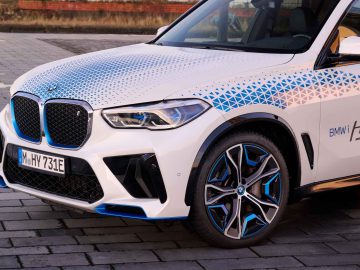 Close-up van de voorkant en zijkant van een witte BMW iX5 Hydrogen SUV met geometrische blauwe details, gemarkeerd met "BMW i Hydrogen NEXT", geparkeerd op een verhard oppervlak.