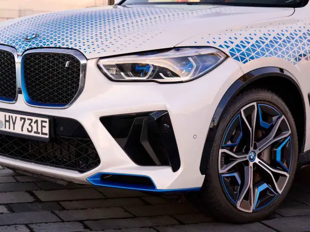 Close-up van de voorste helft van een witte BMW iX5 Hydrogen SUV met blauwe accenten, geometrische patronen op de motorkap en zichtbare kentekenplaat. De auto staat geparkeerd op een geplaveide ondergrond.