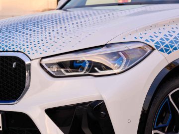 Close-up van de linkervoorzijde van de BMW iX5 Hydrogen, met een witte auto met een geometrisch patroon op de motorkap en een modern koplampontwerp.