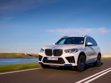 Een witte BMW iX5 Hydrogen SUV glijdt over de weg met een achtergrond van blauwe lucht en bruisend water.