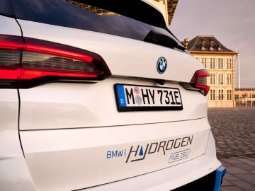 Het achteraanzicht van een witte BMW iX5 Hydrogen met een 'HYDROGEN FUEL CELL'-label op de bumper, Europees kenteken M HY 731E, buiten geparkeerd nabij een groot gebouw.