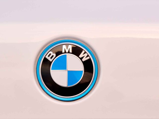 Close-up van een BMW iX5 Hydrogen-logo op een wit oppervlak. Het logo is voorzien van een zwarte buitenring met de letters "BMW" en een blauw-wit geblokte binnencirkel.