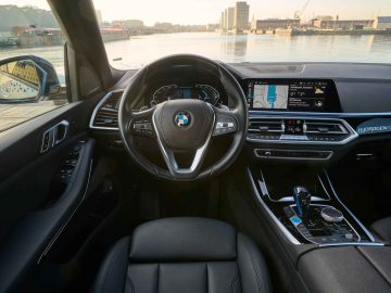 Binnenaanzicht van een BMW iX5 Hydrogen-auto met het stuur, het dashboard met infotainmentscherm en de middenconsole. De auto staat geparkeerd vlakbij een stadsgezicht aan het water dat zichtbaar is door de voorruit.