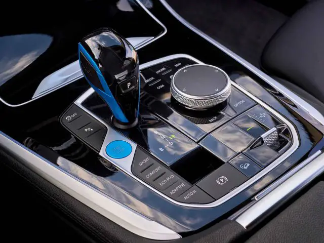 Close-up van het centrale bedieningspaneel van een BMW iX5 Hydrogen met een versnellingspook, verschillende knoppen en een draaiknop. De interface is modern met een strak design in zwart en chromen accenten.