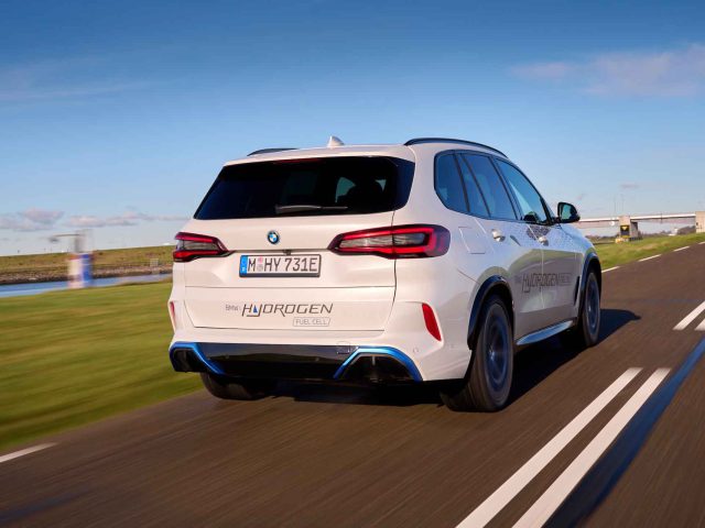 Een witte BMW iX5 waterstofbrandstofcelauto rijdt op een weg geflankeerd door groen gras, met blauwe lucht en wolken op de achtergrond. Op het kenteken staat "M HY 731E.