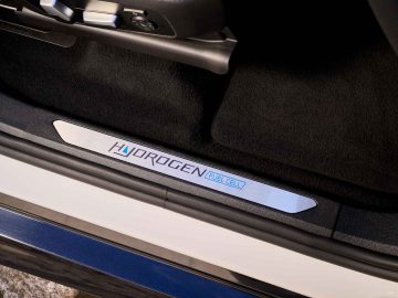 Close-up van de dorpel van een auto met het label 'Hydrogen Fuel Cell', wat aangeeft dat het voertuig waterstofbrandstofceltechnologie gebruikt. Het gebied heeft een metalen en zwarte afwerking, typisch voor BMW iX5 Hydrogen-modellen, en de deur lijkt open te staan.