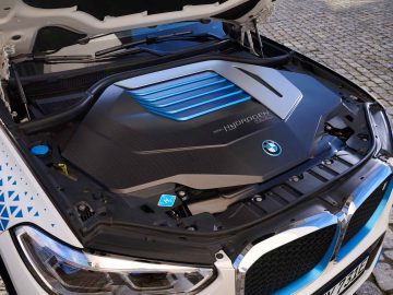 Open motorkap van een auto met daarin een BMW iX5-waterstofmotor met een modern en strak ontwerp, met blauwe en zilveren accenten en een waterstofsymbool op de zwarte kap. Op de achtergrond zijn geplaveide bestrating zichtbaar.