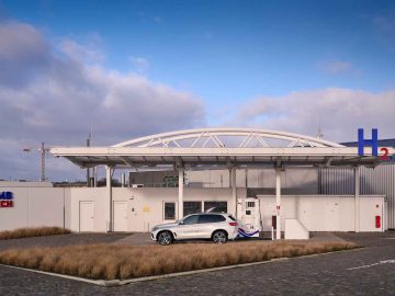 Een wit waterstoftankstation met een witte BMW iX5 Waterstof geparkeerd onder de overkapping. Het tankstation heeft het CMB Tech-logo en staat tegen een gedeeltelijk bewolkte hemel.