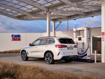 Een witte SUV met het opschrift 'BMW iX5 Hydrogen' tankt bij een waterstofstation onder een gedeeltelijk overdekte overkapping met de tekst 'CMB Tech', waarmee BMW's toewijding aan duurzame innovatie wordt getoond.