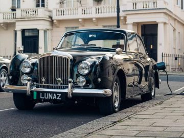 Een klassieke Bentley-auto met een "LUNAZ"-kentekenplaat staat geparkeerd in een stadsstraat, naadloos verbonden met een laadstation voor elektrische voertuigen.