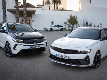 Twee witte auto's met zwarte accenten, waaronder een strakke Opel GSe, staan buiten geparkeerd voor witte gebouwen en palmbomen. De auto aan de linkerkant is een SUV, terwijl de auto aan de rechterkant een sedan is.