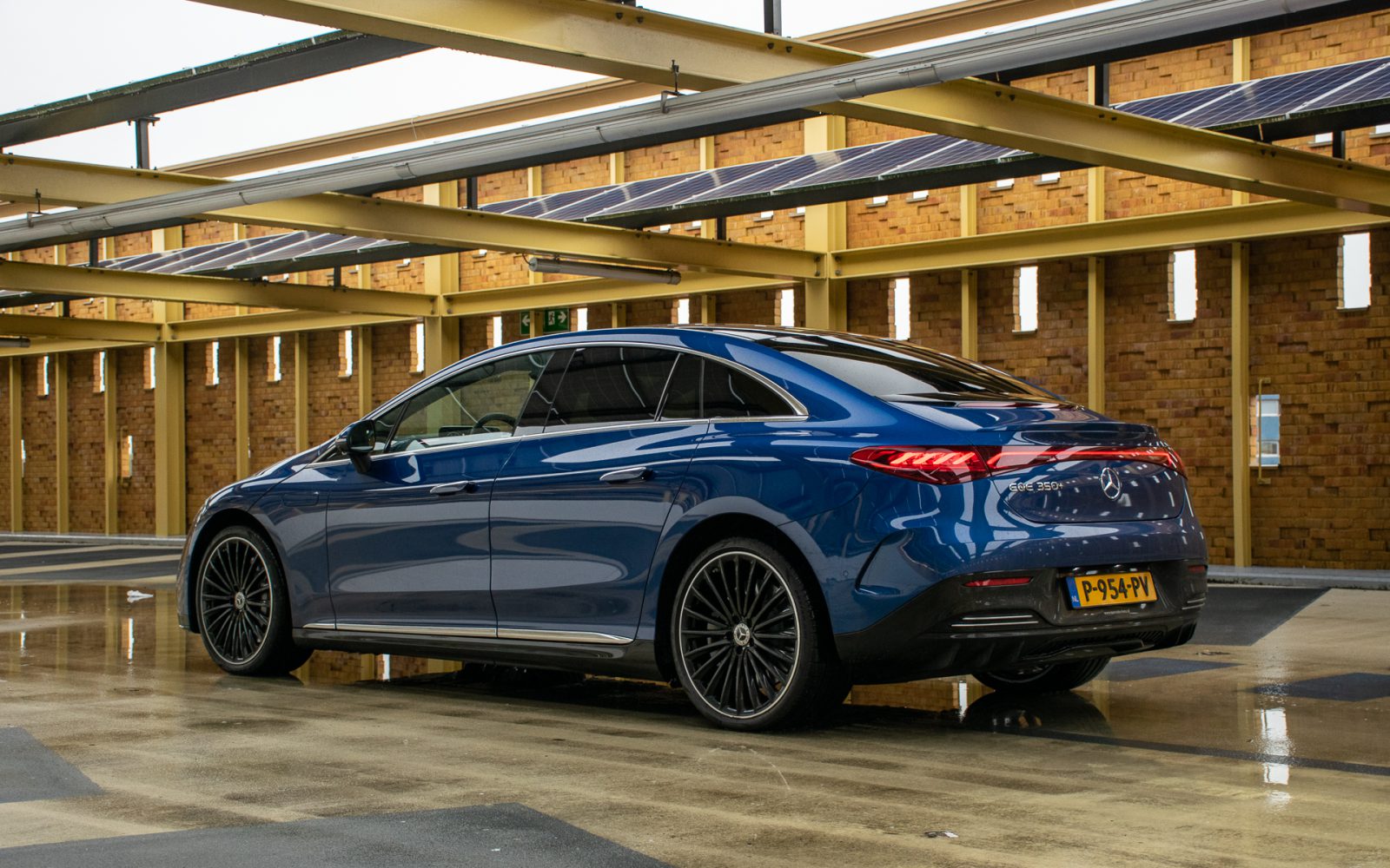 Een blauwe Mercedes-Benz EQE luxe sedan staat geparkeerd onder een constructie met zonnepanelen op het dak. De auto heeft een strak design met stijlvolle velgen en staat op een gladde, natte ondergrond.
