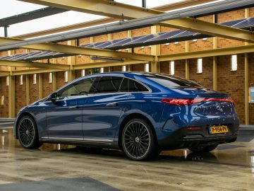Een blauwe Mercedes-Benz EQE luxe sedan staat geparkeerd onder een constructie met zonnepanelen op het dak. De auto heeft een strak design met stijlvolle velgen en staat op een gladde, natte ondergrond.